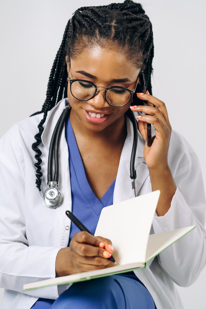 Black female doctor writing on a notebook while talking on a phone, wearing eyeglasses.