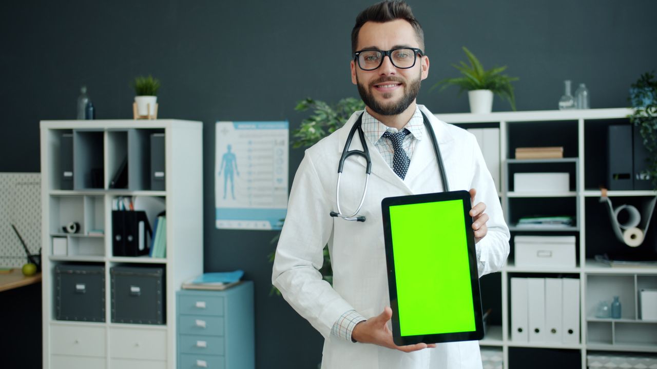 Smiling doctor with a stethoscope presenting a green screen tablet in a modern office environment.