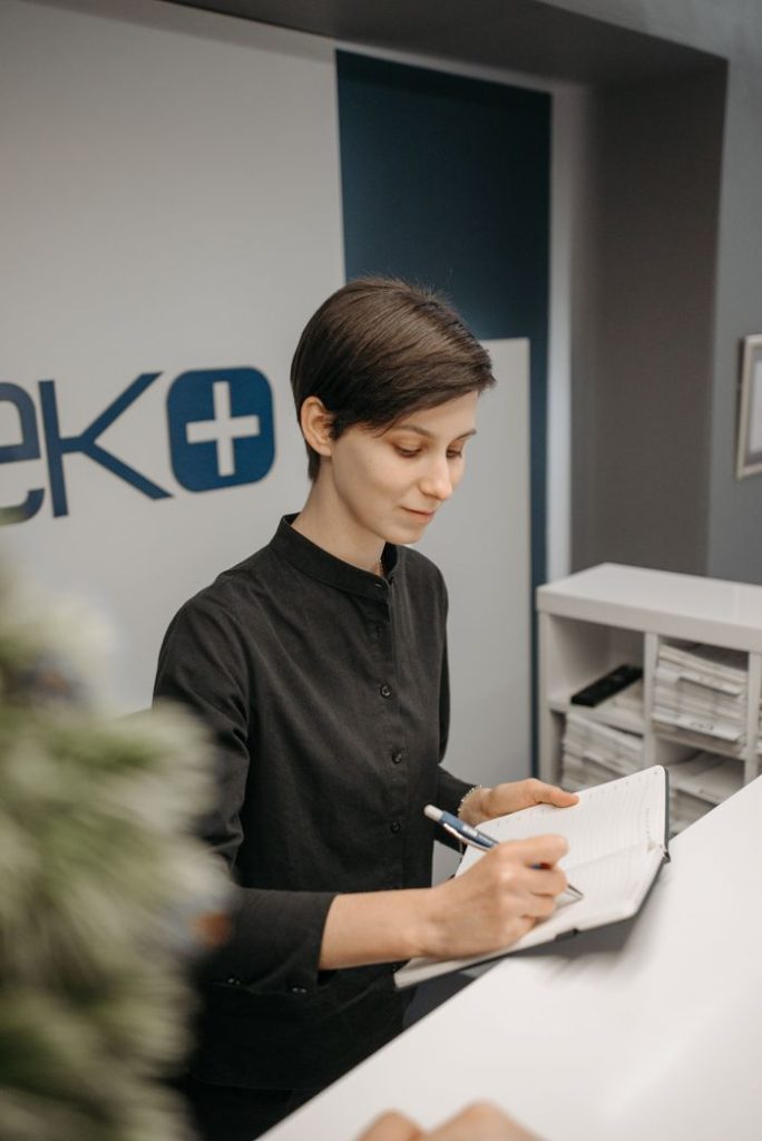Female receptionist writing in a notebook at a clinic reception area, showcasing professionalism.